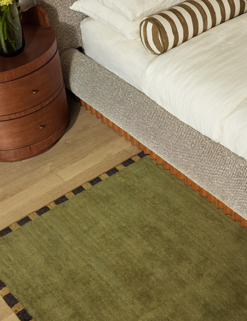May include: A bedroom scene featuring a bed with a patterned headboard and a green rug with a black and gold border. A wooden nightstand with drawers is visible, along with a striped bolster pillow.