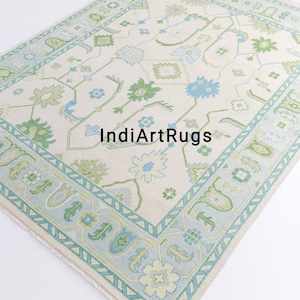 May include: A cream-colored rug with a blue and green floral pattern. The rug has a border with a geometric design. The text "IndiArtRugs" is visible in the center of the rug.