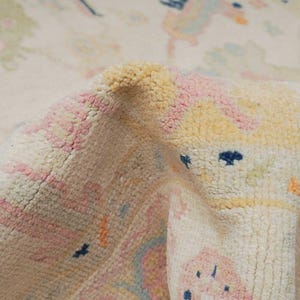 May include: Close-up of a cream-colored rug with a pink, yellow, and blue abstract pattern. The rug has a soft, fluffy texture.
