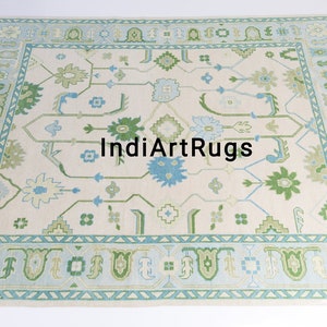May include: A cream-colored rug with a blue and green geometric pattern. The rug has a border with a repeating pattern of triangles and arches. The text "IndiArtRugs" is in the center of the rug.