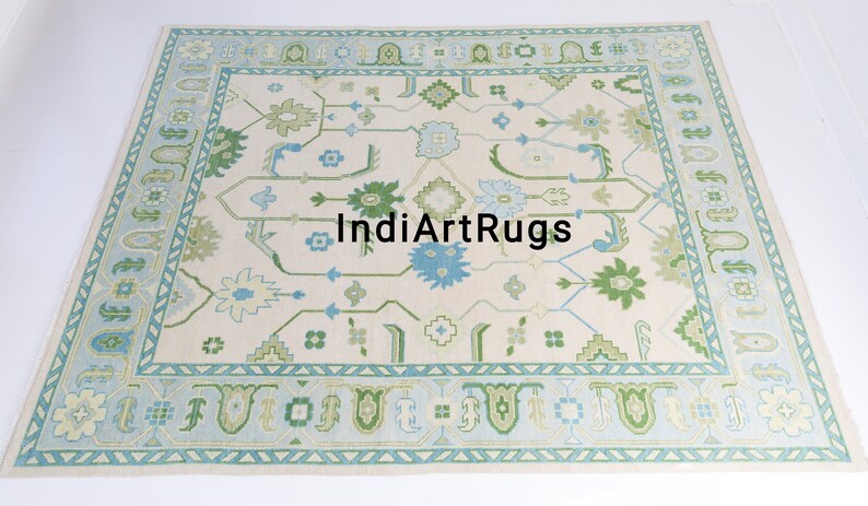 May include: A cream-colored rug with a blue and green geometric pattern. The rug has a border with a repeating pattern of triangles and arches. The text "IndiArtRugs" is in the center of the rug.