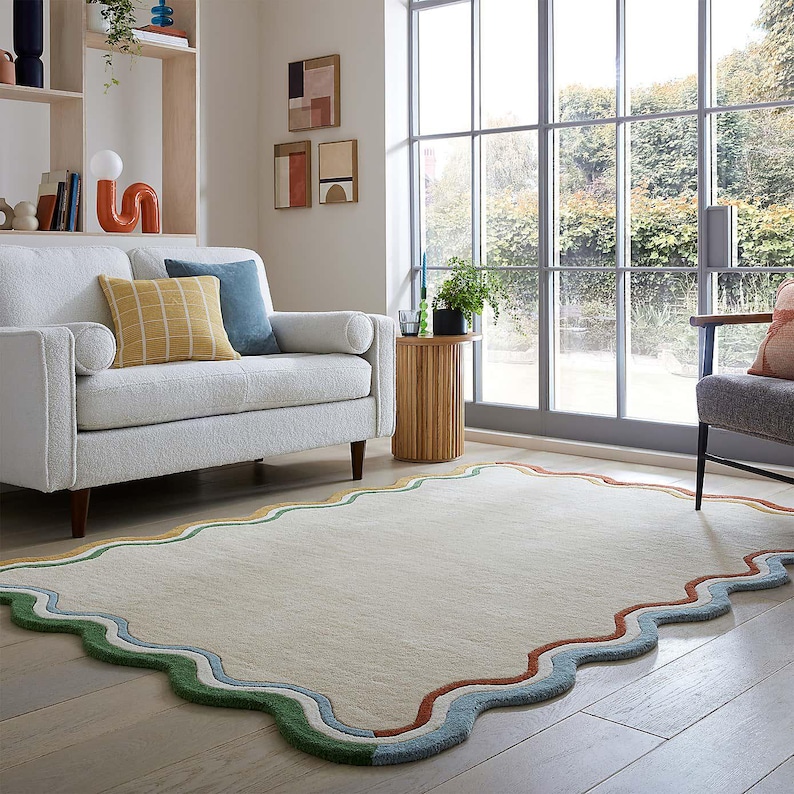 May include: A cream-colored rectangular rug with a wavy, multi-colored border. The border features green, yellow, blue, and red stripes. The rug is placed on a light-colored wooden floor in a living room setting.