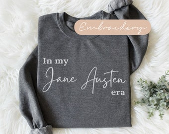 Puede incluir: Una sudadera gris oscuro con el texto "In my Jane Austen era" bordado en blanco en la parte delantera.