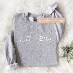 May include: A heather gray sweatshirt with the text "EST. 1994 LIMITED EDITION" in white. The sweatshirt has a crew neck and long sleeves with rolled cuffs. The word "Embroidery" is written on a tan label.