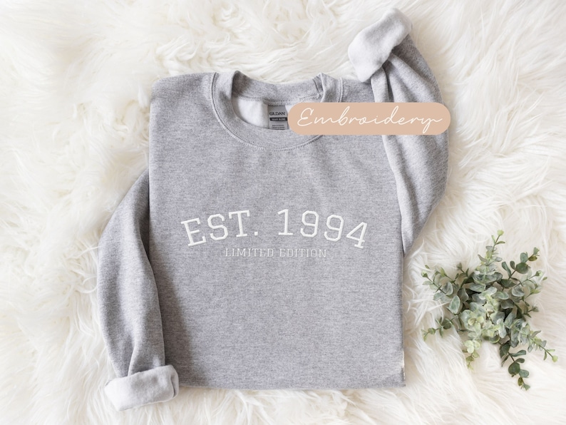 May include: A heather gray sweatshirt with the text "EST. 1994 LIMITED EDITION" in white. The sweatshirt has a crew neck and long sleeves with rolled cuffs. The word "Embroidery" is written on a tan label.