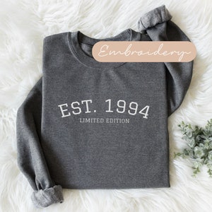 May include: Dark gray sweatshirt with the text "EST. 1994 LIMITED EDITION" in white. The sweatshirt has a crew neck and cuffed sleeves. The background is a white, fluffy surface. A small sprig of greenery is in the lower right corner.