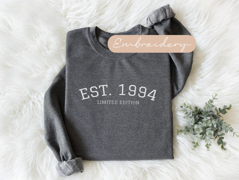 May include: Dark gray sweatshirt with the text "EST. 1994 LIMITED EDITION" in white. The sweatshirt has a crew neck and cuffed sleeves. The background is a white, fluffy surface. A small sprig of greenery is in the lower right corner.