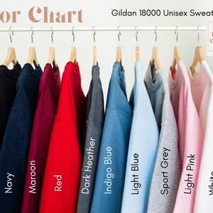 May include: A color chart displays Gildan 18000 unisex sweatshirts in various colors. The sweatshirts are hanging on a white rack. Colors include black, navy, maroon, red, dark heather, indigo blue, light blue, sport grey, light pink, and white.