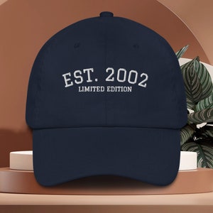 May include: Navy blue baseball cap with white embroidered text reading "EST. 2002 LIMITED EDITION." The cap has a curved brim and a structured crown. The hat is displayed on a neutral background.