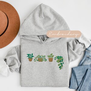 May include: A gray hooded sweatshirt with a design of five potted plants embroidered on the front. The text "Embroidery" is written in a light pink font on a light brown rectangle above the design.