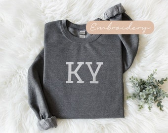 May include: Gray sweatshirt with white embroidered letters "KY" on the front. The sweatshirt is on a white fluffy surface.