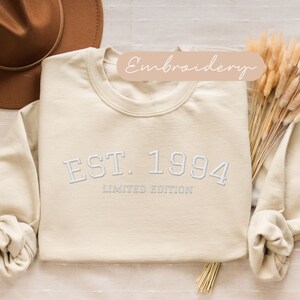 May include: Cream-colored sweatshirt with the embroidered text "EST. 1994 LIMITED EDITION" in white. The long-sleeved top is folded, with a brown hat and dried flowers in the background. The word "Embroidery" is written on a light brown banner.