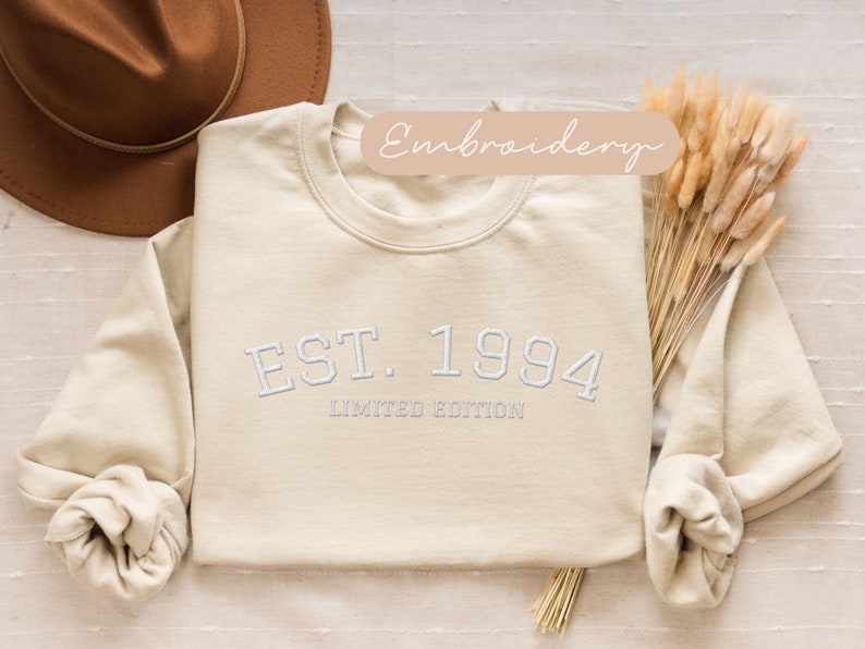 May include: Cream-colored sweatshirt with the embroidered text "EST. 1994 LIMITED EDITION" in white. The long-sleeved top is folded, with a brown hat and dried flowers in the background. The word "Embroidery" is written on a light brown banner.