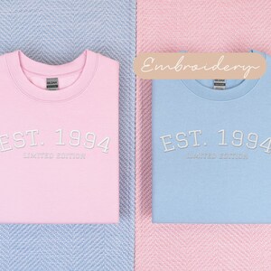 May include: Two sweatshirts, one pink and one light blue, are displayed. Each has the embroidered text "EST. 1994 LIMITED EDITION" in white. The sweatshirts are folded and placed on a blue and pink patterned surface.