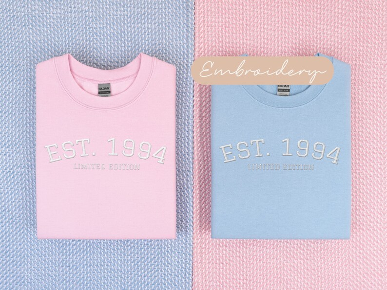 May include: Two sweatshirts, one pink and one light blue, are displayed. Each has the embroidered text "EST. 1994 LIMITED EDITION" in white. The sweatshirts are folded and placed on a blue and pink patterned surface.