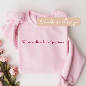 May include: A light pink sweatshirt with the phrase "What excellent boiled potatoes" in a burgundy font. The word "Embroidery" is written in a cursive font on a light brown banner. Pink flowers are in the lower left corner.