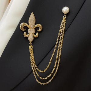 Pode incluir: Um broche de flor-de-lis em tom dourado com cristais pavé transparentes, ligado por três correntes douradas drapeadas a um pino de pérola sintética branca. O broche está preso a uma peça de vestuário de tecido preto.