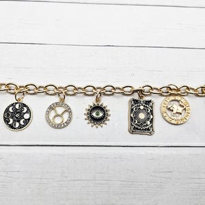 May include: Gold-tone charm bracelet featuring various zodiac and celestial-themed pendants. Charms include a Taurus symbol, a moon phase design, and a sun with an eye. The bracelet has a toggle clasp and is displayed on a white wooden surface.