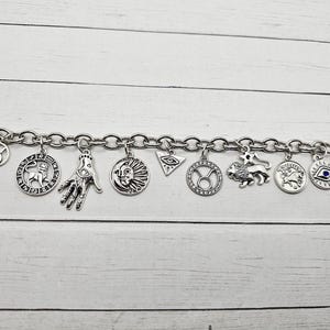 May include: Silver-toned charm bracelet featuring various zodiac symbols, a hand with an eye, a sun face, and an evil eye charm. The bracelet has a toggle clasp and is displayed on a white wooden surface.