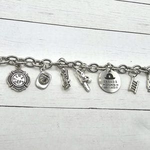 Può includere: Un braccialetto con ciondoli in argento con vari ciondoli a tema pompiere, tra cui un idrante, un casco da pompiere, una scala, un estintore e un ciondolo che dice "Ho baciato un pompiere e mi è piaciuto".