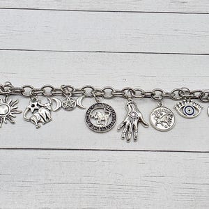 May include: Silver-toned charm bracelet featuring various celestial and zodiac symbols. Charms include a sun, moon, stars, hand, and zodiac signs. The bracelet has a toggle clasp and a blue evil eye charm.