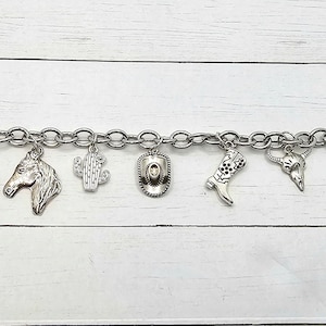 Op de afbeelding: Een zilveren armband met western-thema charms, waaronder een paardenkop, cactus, cowboyhoed, laars, schedel en pijl en boog.