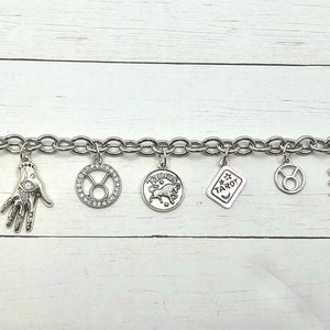 May include: A silver charm bracelet with various charms, including a sun, an eye, a hand, a tarot card, and a Taurus zodiac sign. The bracelet is on a white wooden background.