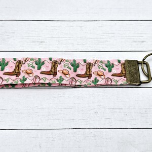 May include: Pink and green key chain with a pattern of cowboy boots and hats. The key chain has a metal ring and a metal clasp.