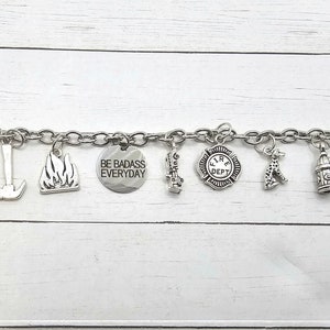 Op de afbeelding: Een zilveren armband met verschillende brandweer-charms, waaronder een brandkraan, een bijl, een brandweerwagen en een brandweerhelm. De armband bevat ook een charm met de tekst "BE BADASS EVERYDAY".