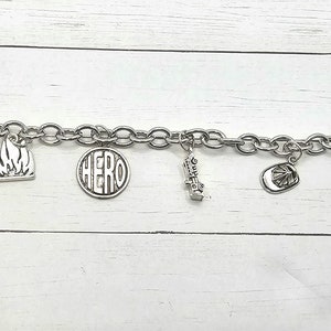Può includere: Un braccialetto con ciondoli in argento con diversi ciondoli, tra cui un logo del corpo dei vigili del fuoco, una fiamma, la parola "HERO", un idrante, un cappello da pompiere e uno spegnimento.