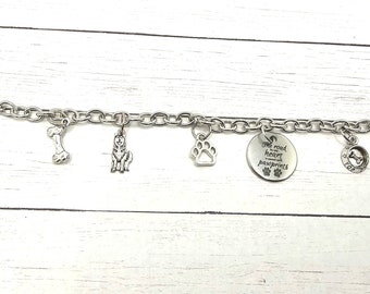 May include: Silver charm bracelet with dog-themed charms, including a bone, paw print, dog silhouette, and a charm with the text "One good heart is worth more than a thousand paw prints".