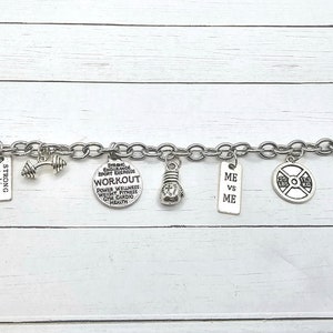May include: A silver charm bracelet with various fitness-themed charms, including dumbbells, a boxing glove, and a weight plate. The charms have text on them, including "Strong not skinny", "Workout", and "Me vs Me".