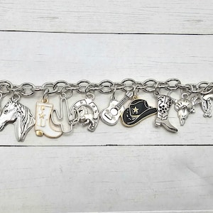May include: A silver charm bracelet with various western-themed charms, including a cactus, horseshoe, cowboy hat, boot, and guitar. The charms are attached to a silver chain bracelet.