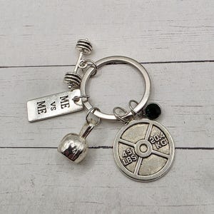 May include: Silver-toned keychain featuring a barbell, dumbbell, and weight plate charm. The weight plate is marked with "45 LBS" and "20.4 KG". A small tag reads "ME VS ME". The keychain is on a silver ring.