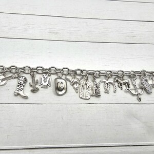May include: A silver charm bracelet with a variety of western-themed charms, including a cowboy boot, a cactus, a horseshoe, a guitar, and a cowboy hat.