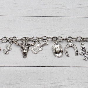 May include: Silver-toned charm bracelet featuring Western-themed charms. Charms include a cowboy boot, guitar, horseshoe, bull skull, and a cross. The bracelet has a toggle clasp and a small blue gem.