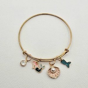 May include: Gold bangle bracelet with charms including a mermaid, a seashell with a pearl, and a fish tail.