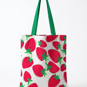 Peut inclure: Un sac fourre-tout blanc orné d'un motif répétitif de fraises rouges avec des feuilles vertes et des graines noires. Le sac est doté de deux longues anses en tissu vert uni fixées sur le dessus, offrant une prise solide pour le transport.