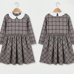 May include: Two gray plaid dresses with long sleeves and white Peter Pan collars hang on wooden hangers. The dresses have a red plaid pattern and a gathered skirt. The dresses are displayed against a plain white background.