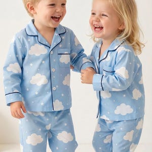 Peut inclure: Deux enfants portant des pyjamas bleus clairs assortis avec un motif de nuages et d'étoiles. Les pyjamas ont une chemise boutonnée avec une poche et un pantalon long. La bordure est bleu marine.