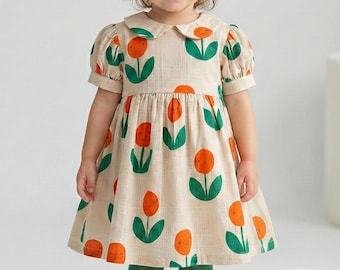 Puede incluir: Una niña pequeña con un vestido beige con estampado floral naranja y verde. El vestido tiene cuello Peter Pan y mangas cortas. La niña también lleva medias verdes, zapatos naranjas y un lazo naranja en el pelo.