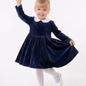 May include: A navy blue velvet dress with a white collar and long sleeves. The dress has a flared skirt and is paired with white tights and white Mary Jane shoes. A small white flower is in the child's hair.