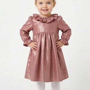 May include: A little girl in a dusty rose dress with a ruffled collar and long sleeves. The dress has a metallic sheen. She wears white tights and matching dusty rose Mary Jane shoes with a bow. Her hair is styled with a matching bow.