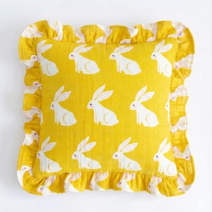 May include: Square yellow pillow with a white rabbit pattern. The pillow has a ruffled edge with a repeating pattern of rabbits and a yellow background. The rabbits have pink ears and noses.