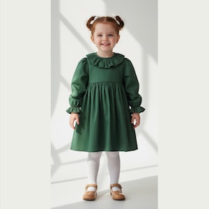 May include: A young child in a forest green dress with a frilled collar and cuffs. The dress has a gathered skirt. The child is also wearing white tights and brown Mary Jane shoes. The background is a plain white wall.