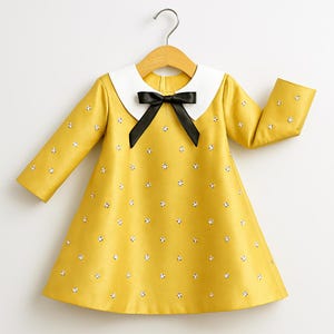 May include: A yellow dress with long sleeves, a white collar, and a black bow. The dress is adorned with small, white floral accents. The dress is hanging on a wooden hanger.