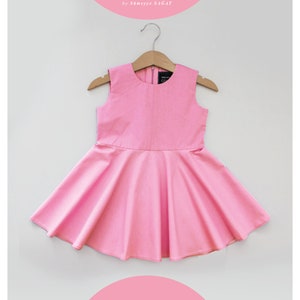 Puede incluir: Un vestido rosa sin mangas con cuello redondo y falda acampanada. El vestido está colgado en una percha de madera. El vestido está etiquetado con "Lucy Rise Kids Wear".