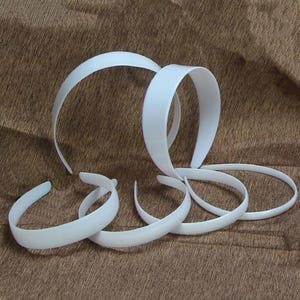 Pode incluir: Várias tiaras de plástico branco de diferentes formas e tamanhos estão dispostas sobre uma superfície marrom. As tiaras são curvas e lisas, com um acabamento brilhante. Algumas são circulares, outras têm um design mais alongado e curvo.