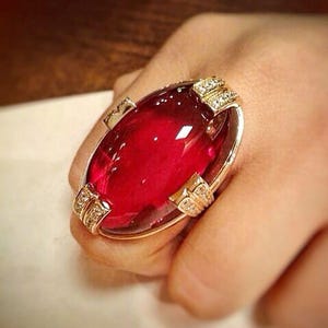 Puede incluir: Un anillo dorado con una gran gema roja oscura de forma ovalada. El anillo tiene gemas claras más pequeñas engastadas en engastes dorados decorativos. Una pieza de joyería audaz.