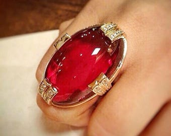 May include: A gold-toned ring featuring a large, oval-shaped, deep red gemstone. The ring is accented with smaller, clear gemstones set in decorative gold settings on the sides. The ring is a statement piece of jewelry.